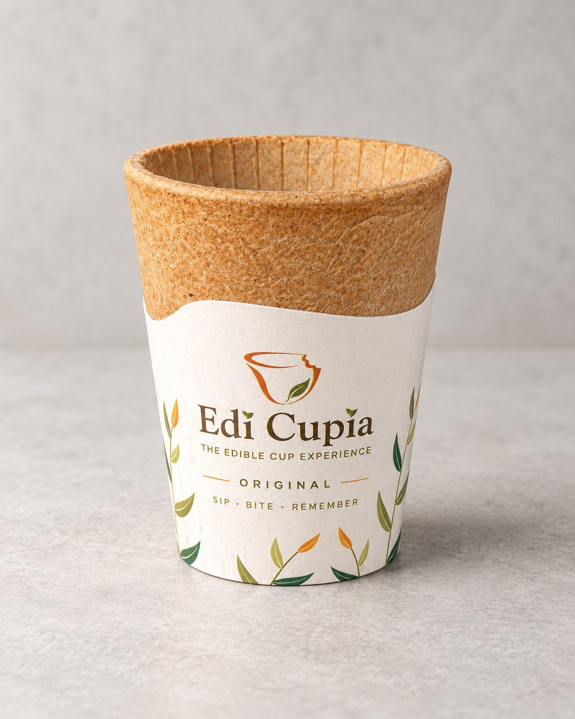 Une tasse comestible en gaufrette Edi Cupia Original dans son manchon ivoire à motif de feuilles, sur une surface de pierre gris pâle.